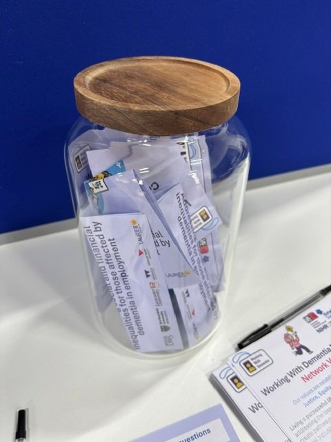 A large jar is on a table with papers inside that people have written on