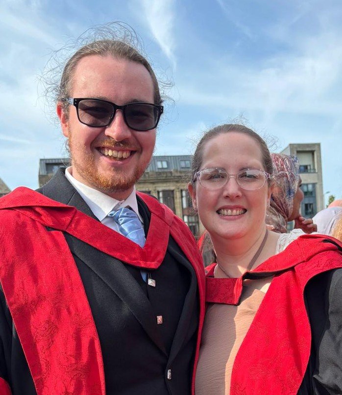 Connor and Rachel are looking at the camera and smiling, wearing their black and red graduation robes.