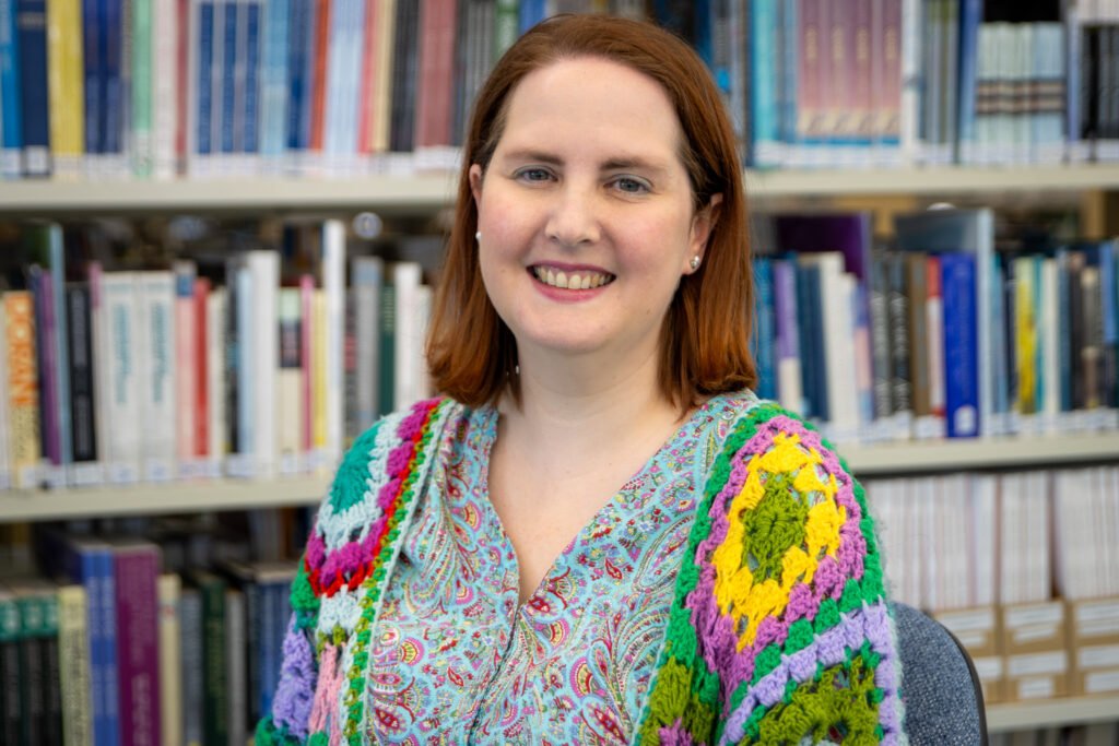 Rachel is wearing a crochet cardigan and is smiling at the camera with books behind her.