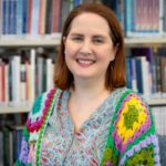 Rachel is wearing a crochet cardigan and is smiling at the camera with books behind her.